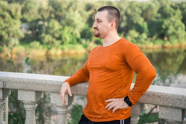 Fototapeta A man in workout clothes rests on a railing with a scenic view
