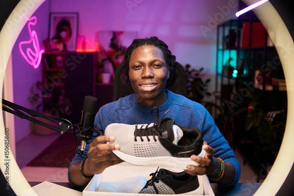 Fototapeta Portrait of Black man showing running shoes in detailed studio setup with plants and neon lights, displaying content creating tools in professional environment