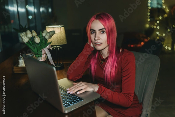 Fototapeta A girl with red hair and a red dress is sitting at a laptop in a cafe