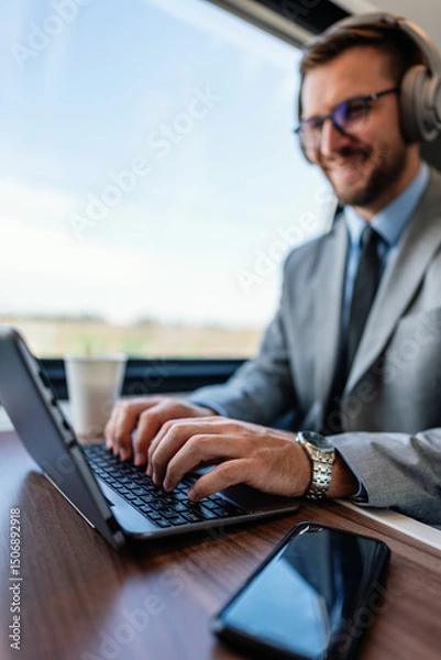 Obraz Young businessman wearing headphones and suit is making a video conference on his laptop while enjoying a cup of coffee during his train journey