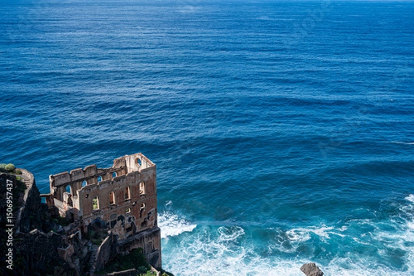 Obraz A small building is on a rocky cliff overlooking the ocean. The ocean is calm and blue, with no visible waves