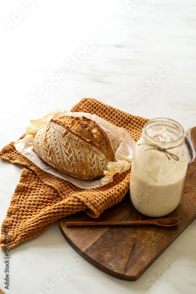 Obraz Crispy sourdough bread sits beside a glass jar filled with active starter, accompanied by a rustic cloth and a wooden board in a warm kitchen atmosphere.