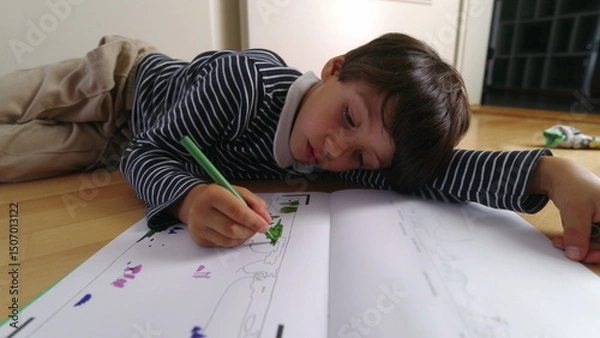 Fototapeta Young boy deeply engaged in drawing with a green pen while lying on the floor, focused on coloring in a book, intense creative concentration, indoor art time