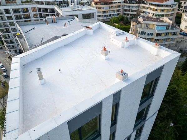 Fototapeta Aerial view of modern residential buildings under construction, featuring flat white rooftops, scaffolding, and unfinished facades. Surrounding neighborhood with completed homes in background