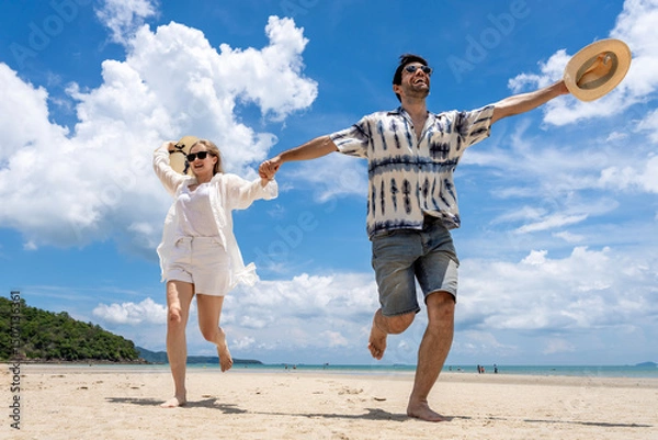 Obraz Couple joyfully running on beach holding hands under sunny sky with clouds in summer afternoon