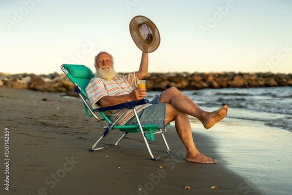 Fototapeta Cheerful elderly man relaxing on the beach, sitting in a chair with a glass of orange juice, smiling and waving his straw hat, enjoying retirement, freedom and happiness during golden sunset.