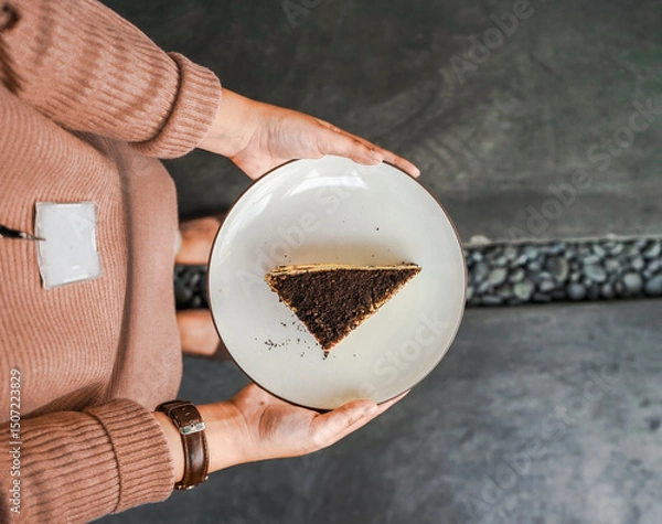 Fototapeta Hands holding a white plate with a tempting piece of layered cake, with chocolate crumbs and cream filling. A delicious dessert