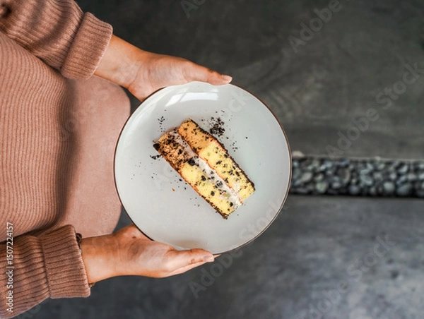 Fototapeta Hands holding a white plate with a tempting piece of layered cake, with chocolate crumbs and cream filling. A delicious dessert