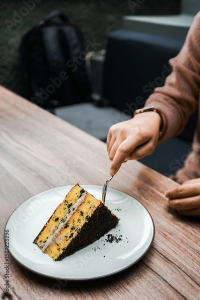 Fototapeta A person cuts into a delicious slice of layered cake with chocolate crumbs and creamy filling, served on a white plate on a wooden table