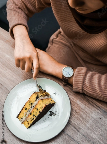 Fototapeta A person cuts into a delicious slice of layered cake with chocolate crumbs and creamy filling, served on a white plate on a wooden table