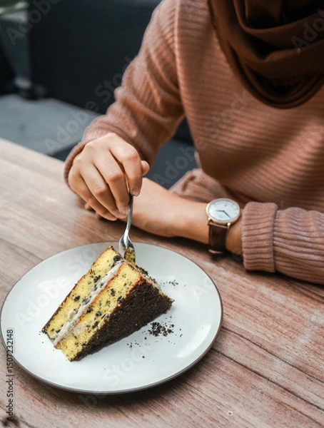 Fototapeta A person cuts into a delicious slice of layered cake with chocolate crumbs and creamy filling, served on a white plate on a wooden table