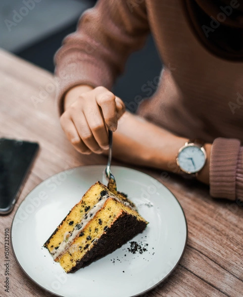 Fototapeta A person cuts into a delicious slice of layered cake with chocolate crumbs and creamy filling, served on a white plate on a wooden table