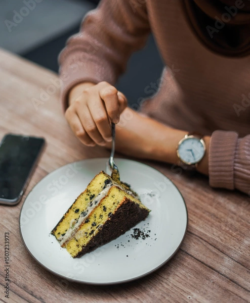 Fototapeta A person cuts into a delicious slice of layered cake with chocolate crumbs and creamy filling, served on a white plate on a wooden table