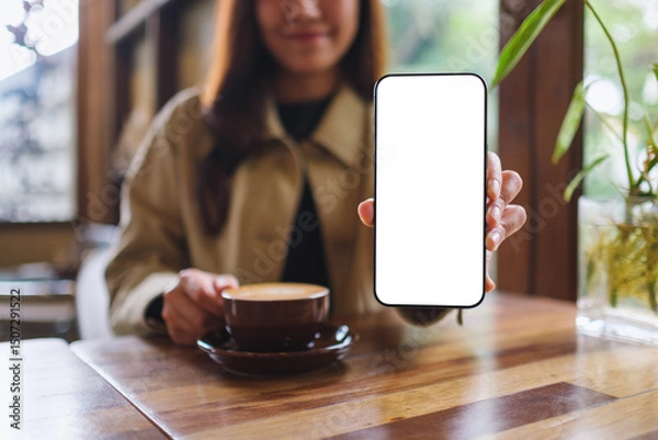 Fototapeta Mockup image of a woman holding and showing mobile phone with blank white screen in cafe