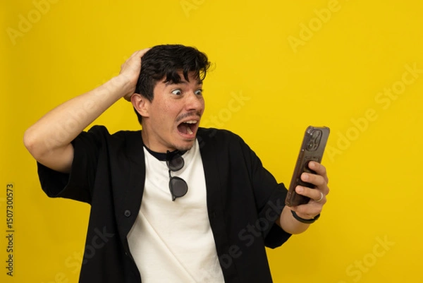 Fototapeta Young freckled man with a mustache with a scared expression while looking at his cell phone on a yellow background with space to write advertising