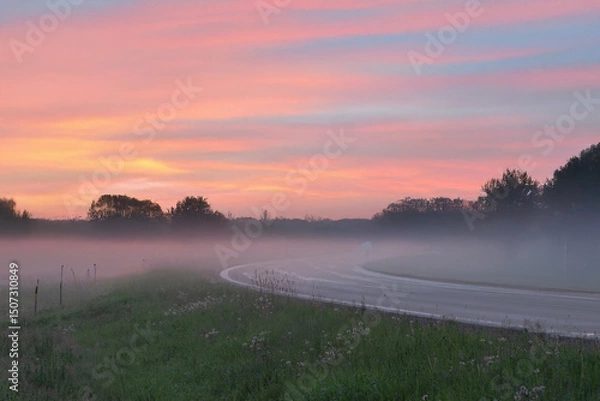 Fototapeta A serene rural road at sunrise with a misty foreground, silhouetted trees, and a pink-orange sky full of scattered clouds.
