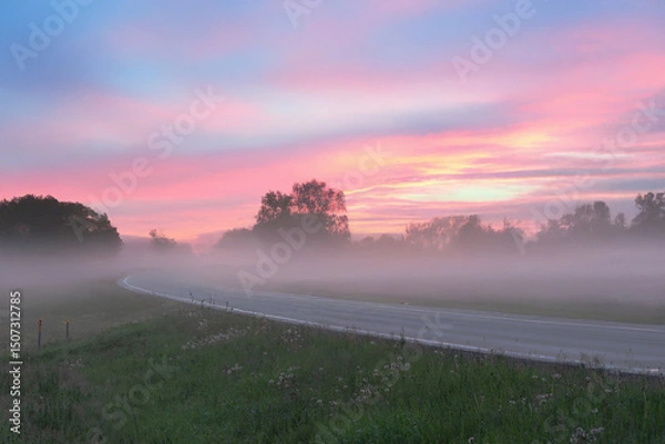 Fototapeta A serene rural road at sunrise with a misty foreground, silhouetted trees, and a pink-orange sky full of scattered clouds.