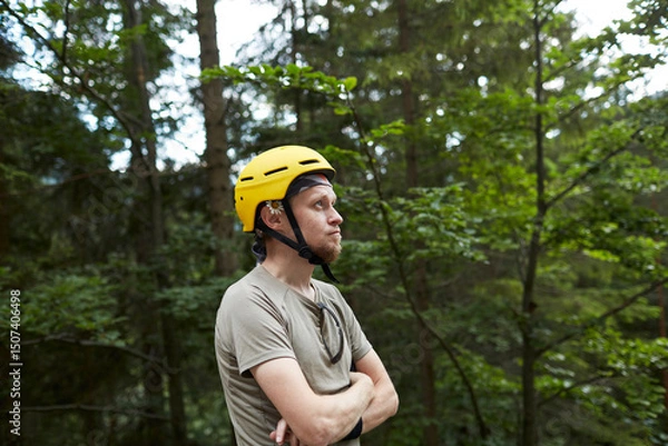 Fototapeta Man wearing a yellow helmet stands in a forest while contemplating nature in the afternoon