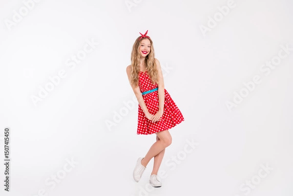 Fototapeta Happy young woman in a red polka-dot dress posing playfully on white, embodying retro charm with stylish details and a joyful attitude.