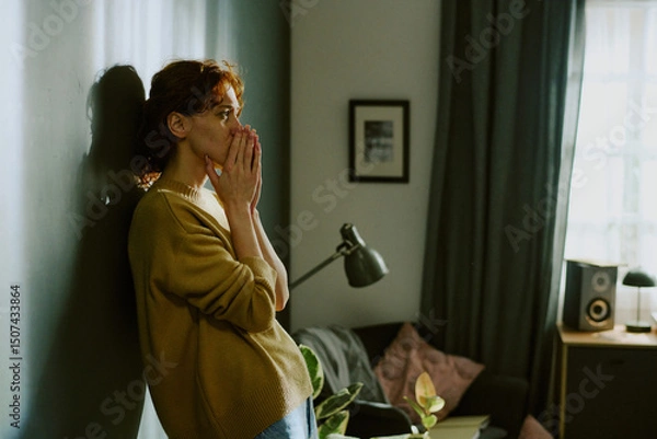 Obraz Woman standing by wall in living room, crying. Room decorated with lamp, framed photo, and few plants, creating a serene and thoughtful atmosphere