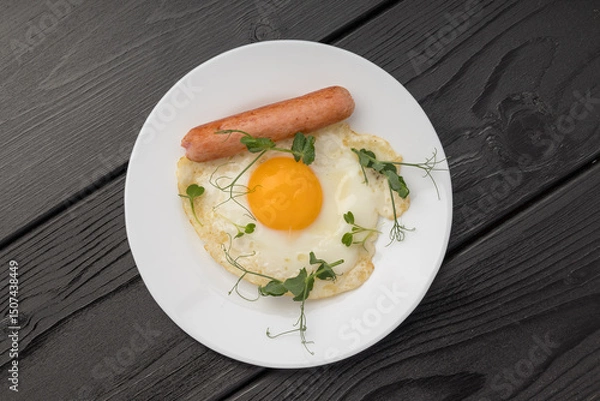 Obraz A fried egg with a sausage and green sprouts on a white plate, dark backdrop.