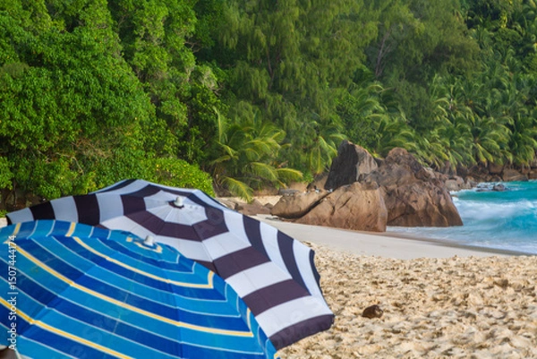 Obraz Umbrella on the sandy beach. Blue Indian Ocean, blue sky with clouds, green rocky hills and palm leaves around, ANSE INTENDANCE, MAHE ISLAND, THE SEYCHELLES