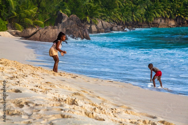 Obraz ANSE INTENDANCE, MAHE, SEYCHELLES - June 2021: children playing in the sand on the beach. Blue water of the Indian Ocean, green rocky hills and a palm in the background