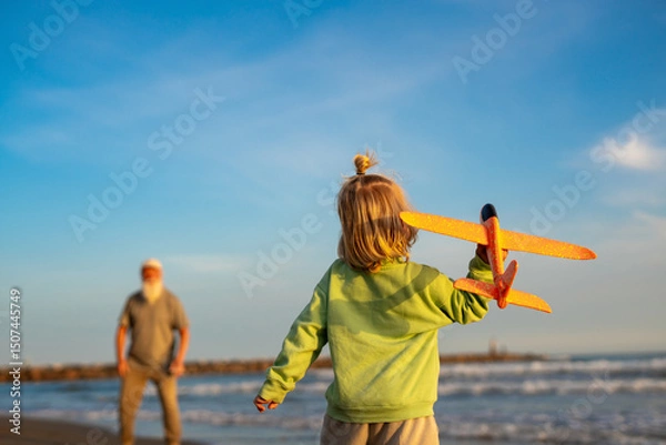Fototapeta Little boy holding an orange toy airplane on the beach while looking at his grandfather in the background, enjoying playful family time by the sea during golden hour.