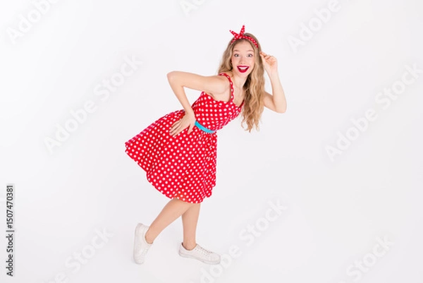 Fototapeta Smiling young woman in retro red polka dot dress posing playfully against a white background, expressing happiness