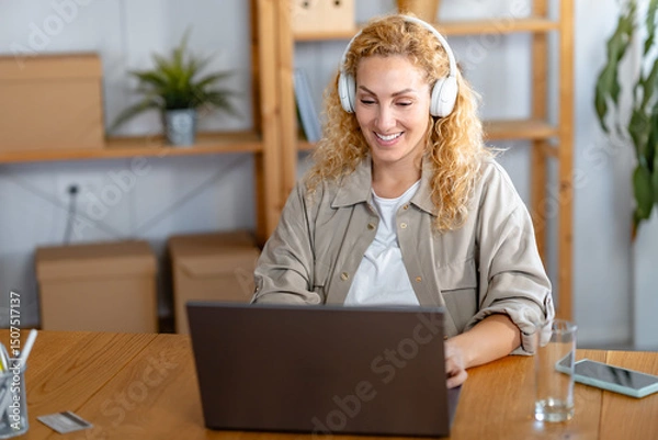 Fototapeta Smiling woman wearing headphones works on her laptop in a cozy home office setting, staying focused and engaged during an online meeting or remote work session.