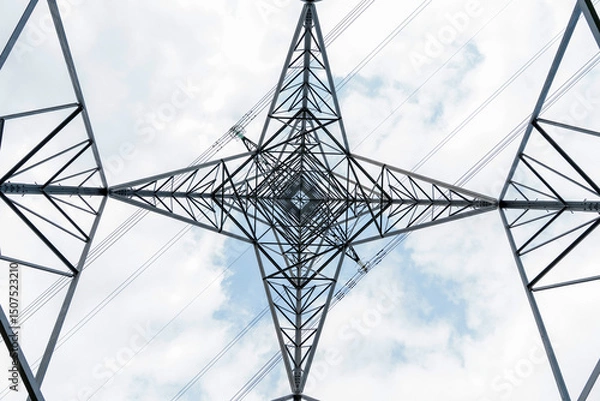 Obraz View of steel power tower from below against a cloudy sky, illustrating symmetrical structure pattern and lines  of energy transmission infrastructure