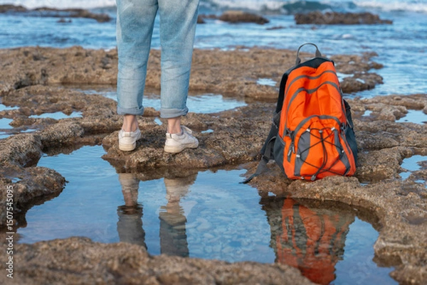 Obraz Backpacker explores rocky shoreline with vibrant orange backpack during daylight hours near the water