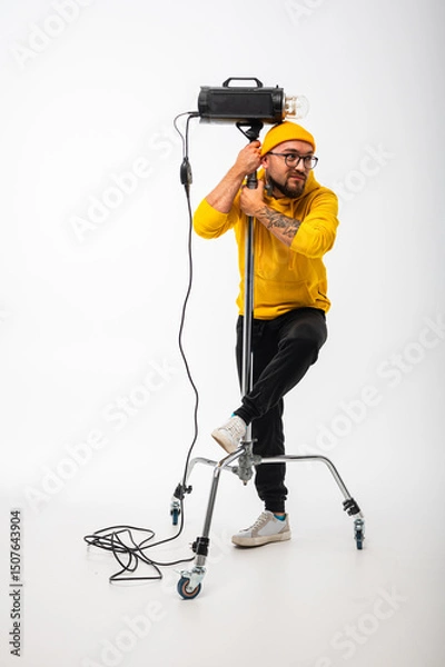 Obraz Portrait of a young man in yellow hoodie and beanie posing with a studio light against white background. Photographer concept, creative professional look