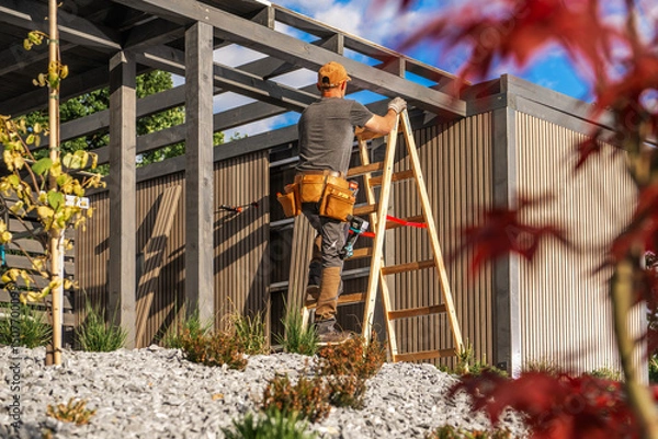 Fototapeta Construction Worker Climbs Ladder to Work on Modern Building in Sunny Outdoor Setting