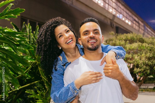 Obraz Young Latin man smiles as his cheerful girlfriend hugs him from behind in a flowery cityscape