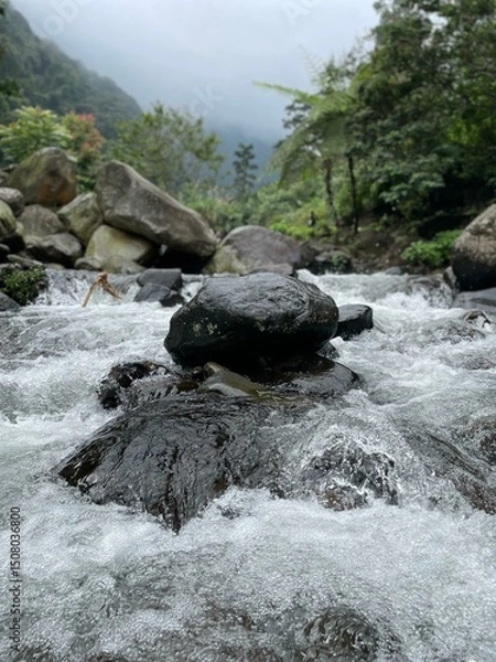 Fototapeta Crystal-clear river flowing over smooth rocks, framed by lush forest and misty mountain views.