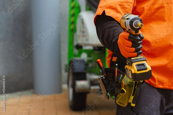 Obraz Close-up of a construction worker with a tool belt, wearing protective gloves, holding a cordless drill. Concept on a construction site with green industrial equipment.