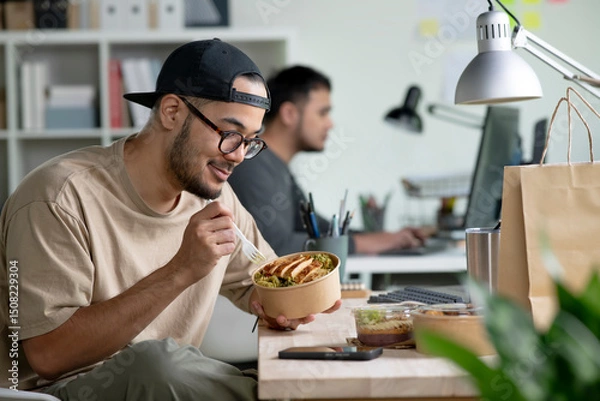 Fototapeta Young Asian man eating healthy meal while working, Businessman having a lunch break at the office