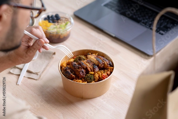 Fototapeta Young Asian man eating healthy meal while working, Businessman having a lunch break at the office, focus on food