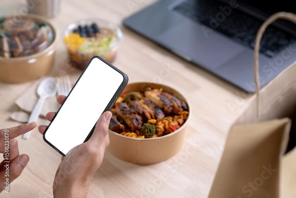 Fototapeta Hand holding a smartphone with blank white screen over paper bowl with healthy food, Young man, woman eating healthy meal while working, Businessperson having a lunch break at the office or home.