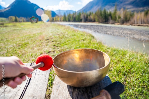 Obraz Point of view of hand playing a Tibetan singing bowl near mountain river in Altai. Concept of personal ritual, sound healing, and spiritual harmony with wild nature.