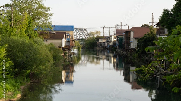 Obraz Canal with Stilt Houses and Reflections, Framed by Greenery and Railway Bridge in the Distance