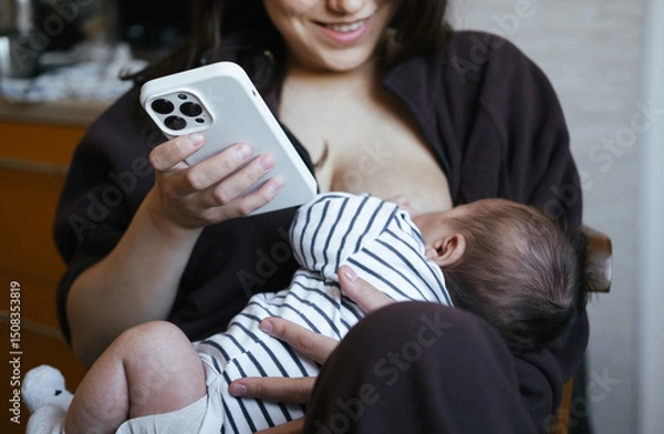 Fototapeta Beautiful young woman is breastfeeding her baby boy at home, close-up view. She is working online at the same time, remote work from home. Chatting with friend, having online shopping. 