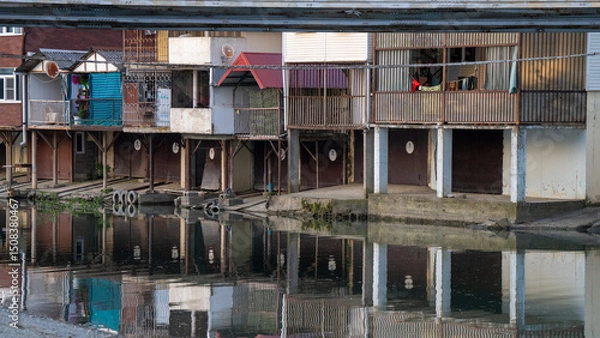 Obraz Riverside Stilt Houses with Reflections on Calm Water in a Rustic Settlement