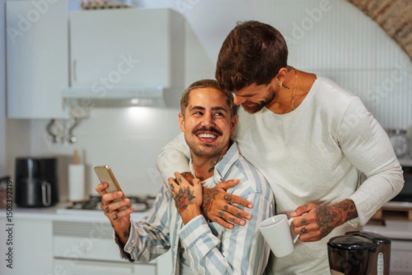 Fototapeta Tattooed gay couple hugging while drinking coffee and using smart phone