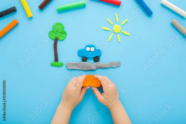 Obraz Little child boy hands making and creating car, tree and sun from colorful modeling clay on light blue table background. Pastel color. Closeup. Point of view shot. Top down view.