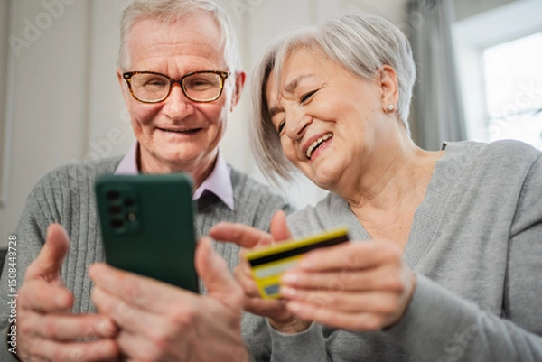 Fototapeta Senior couple man woman shopping online with smartphone paying with credit card. Old people buying on Internet enter credit card details at home indoors. Online shopping delivery service ecommerce