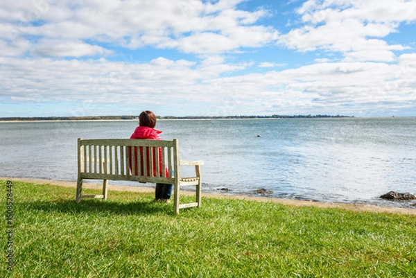 Fototapeta Woman sitting  on a wooden bench in front of a scenic  bay on  a sunny autumn morning