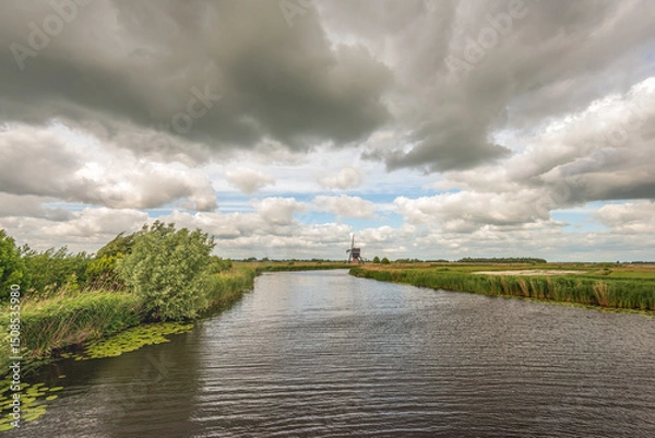 Fototapeta A characteristic Dutch polder landscape featuring a historic thatched hollow post windmill, set beside a wide canal. Captured on a windy spring day under a dramatic sky filled with dark clouds