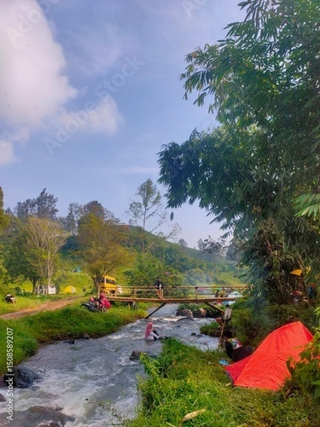 Fototapeta eaceful riverside camping with clear skies, lush greenery, and a wooden bridge over flowing water.